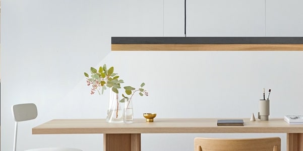 A modern linear pendant light hanging over a light-colored wooden dining table with minimalist decor.